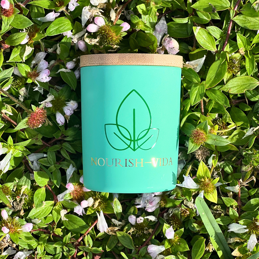 Nourish-Vida natural candle in a matte teal jar with a bamboo lid, featuring a gold leaf logo, resting on green foliage.