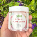 Hand holding a jar of Nourish Glow Peach Body Scrub with green leaves and purple flowers in the background